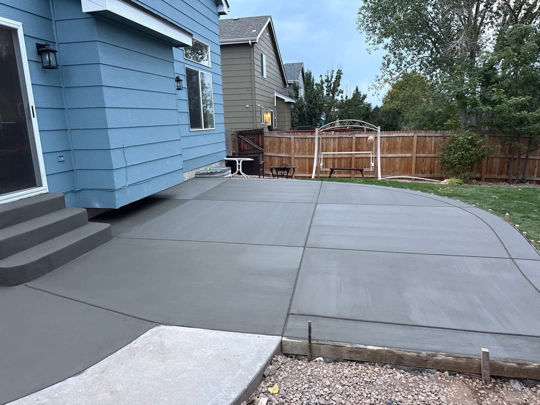 Freshly poured concrete patio beside a house with steps, featuring a backyard swing and fence.