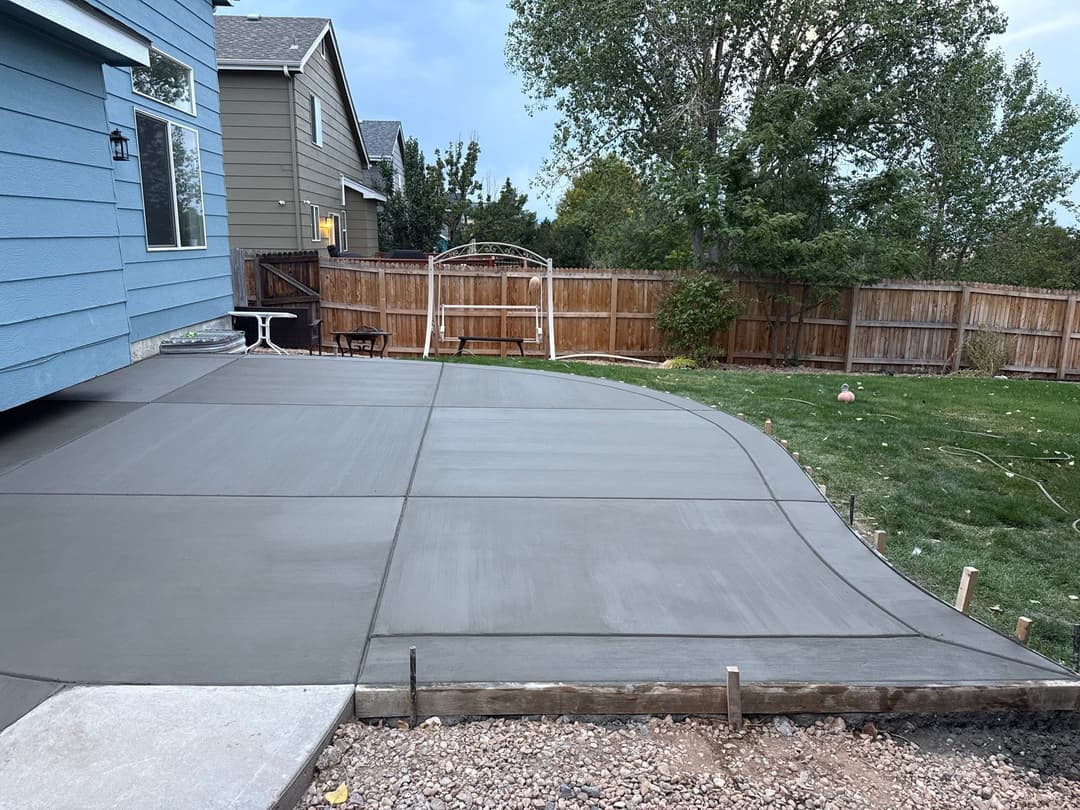 Newly poured concrete patio with curved edge in residential backyard, surrounded by green grass and trees.