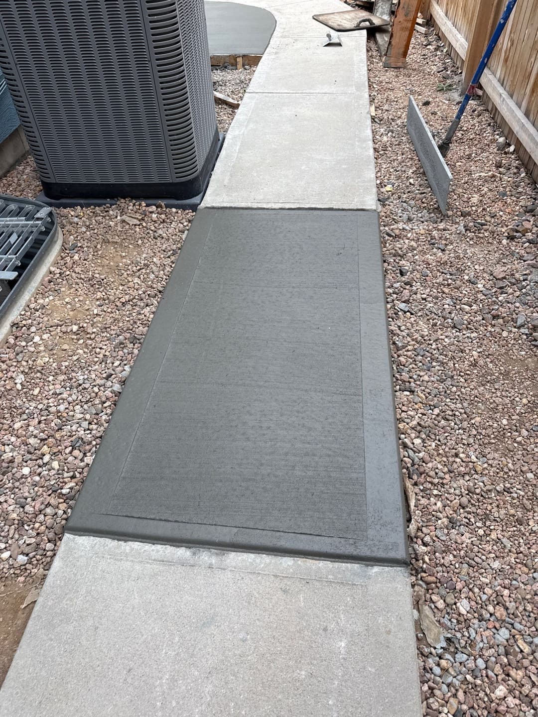 Freshly poured concrete sidewalk with gravel border and equipment in a residential yard.