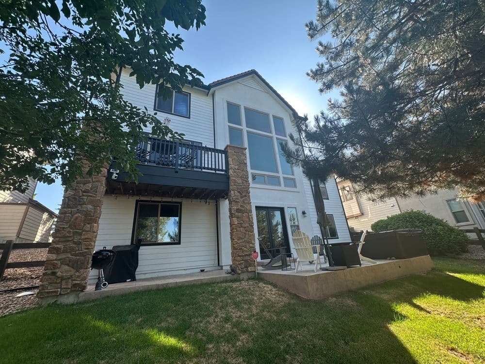 Modern two-story house with large windows, balcony, and outdoor seating area surrounded by greenery.