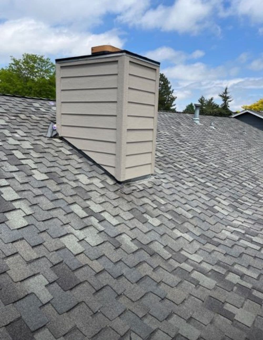 Chimney on shingled roof with blue skies and trees in the background.