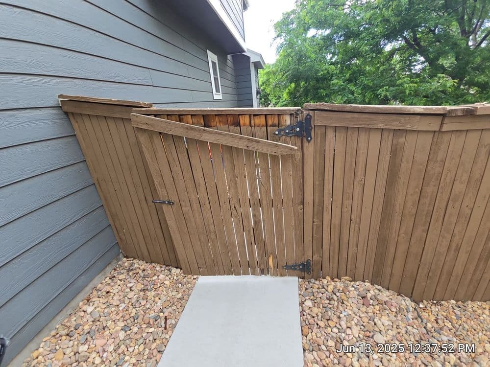 Damaged wooden fence gate leaning against a house with gravel pathway and greenery in background.
